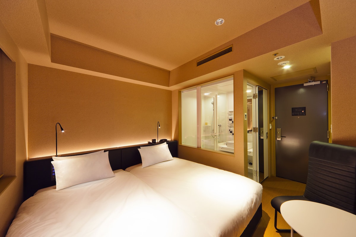 A hotel room with a large bed, bedside lamps, a small table, and a shower area visible through a glass door.