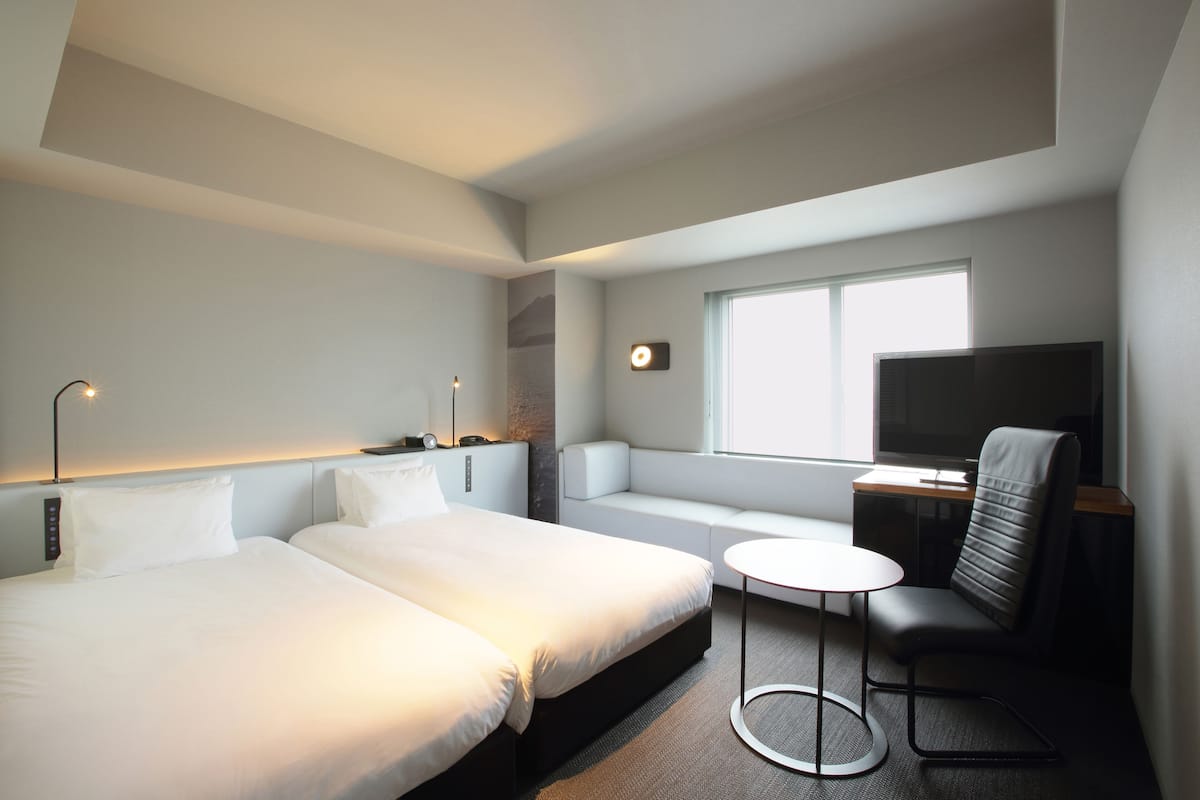 A hotel room with two beds, a small round table, a sofa, a TV, and a window with natural light.