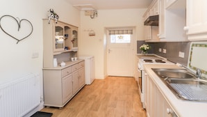 Une cuisine compacte avec des meubles blancs, un petit réfrigérateur, une cuisinière et un évier. Un décor en forme de cœur orne le mur, et une fenêtre offre une vue agréable.