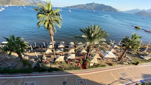 Un hotel frente a la playa con tumbonas y sombrillas, un paseo bordeado de palmeras y vistas al mar y a los barcos.