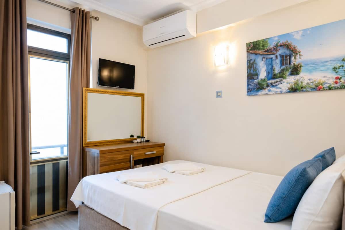 A hotel room with a bed, a TV, a painting, a window with curtains, and an air conditioning unit.