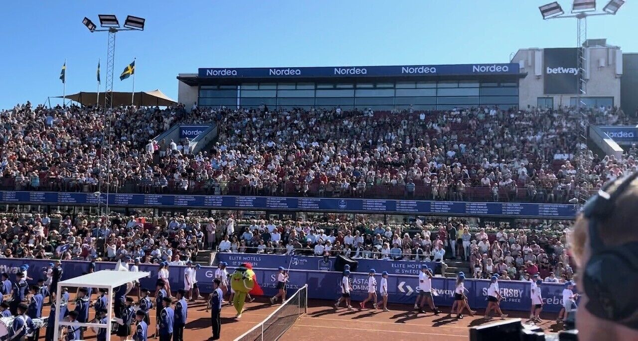 Un estadio de tenis con múltiples niveles de asientos, publicidad de Nordea y Betway, y un animador en la cancha.