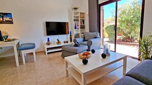 A modern living room with a flat-screen TV, a bookshelf, a dining area, and a large window with a view of greenery. - House CASA MAJA Costa Calma Oceanview Garden WiFi (Costa Calma)