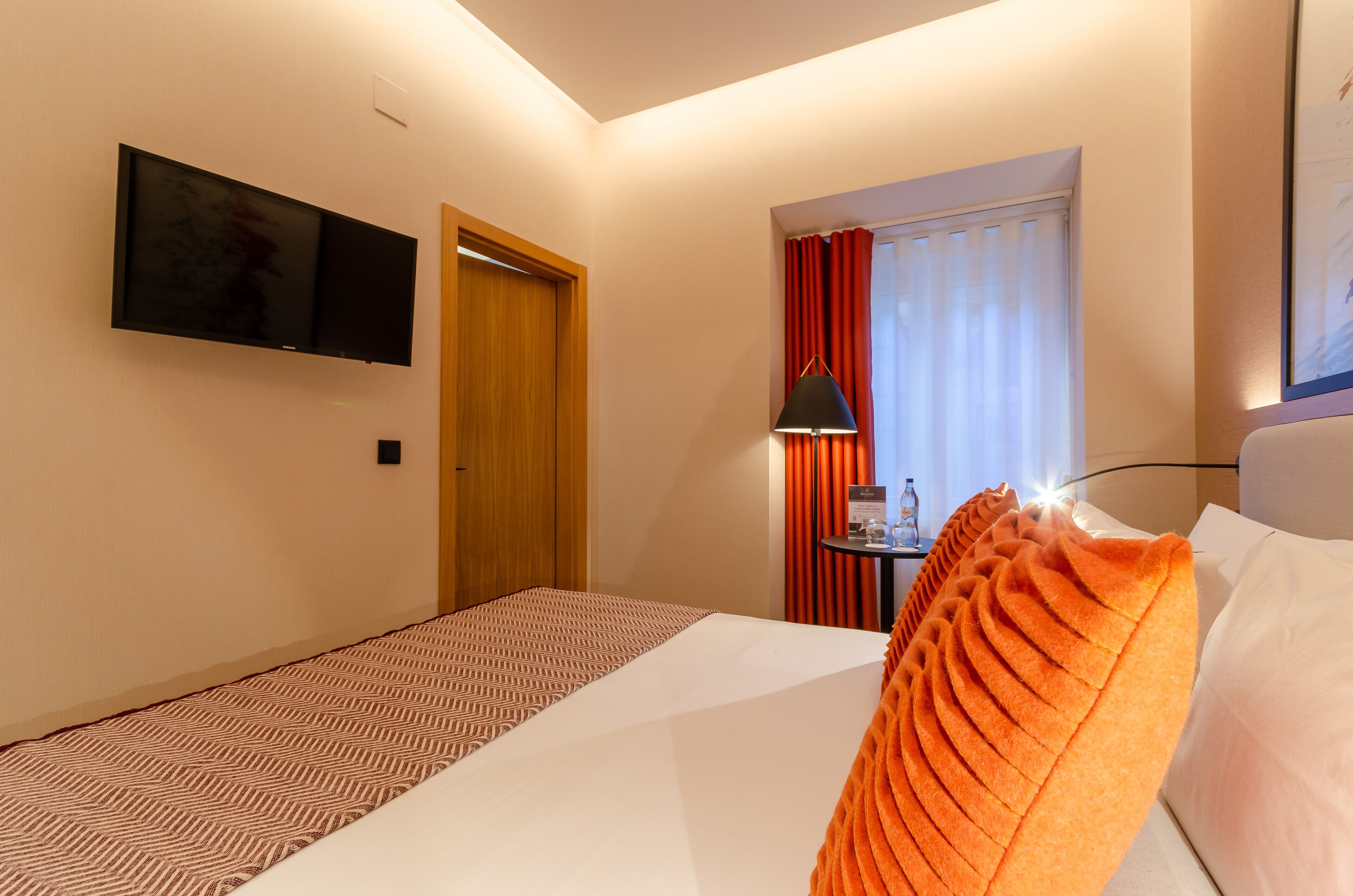 A hotel room with a bed, a TV mounted on the wall, a door, a window with curtains, and a table with a lamp.