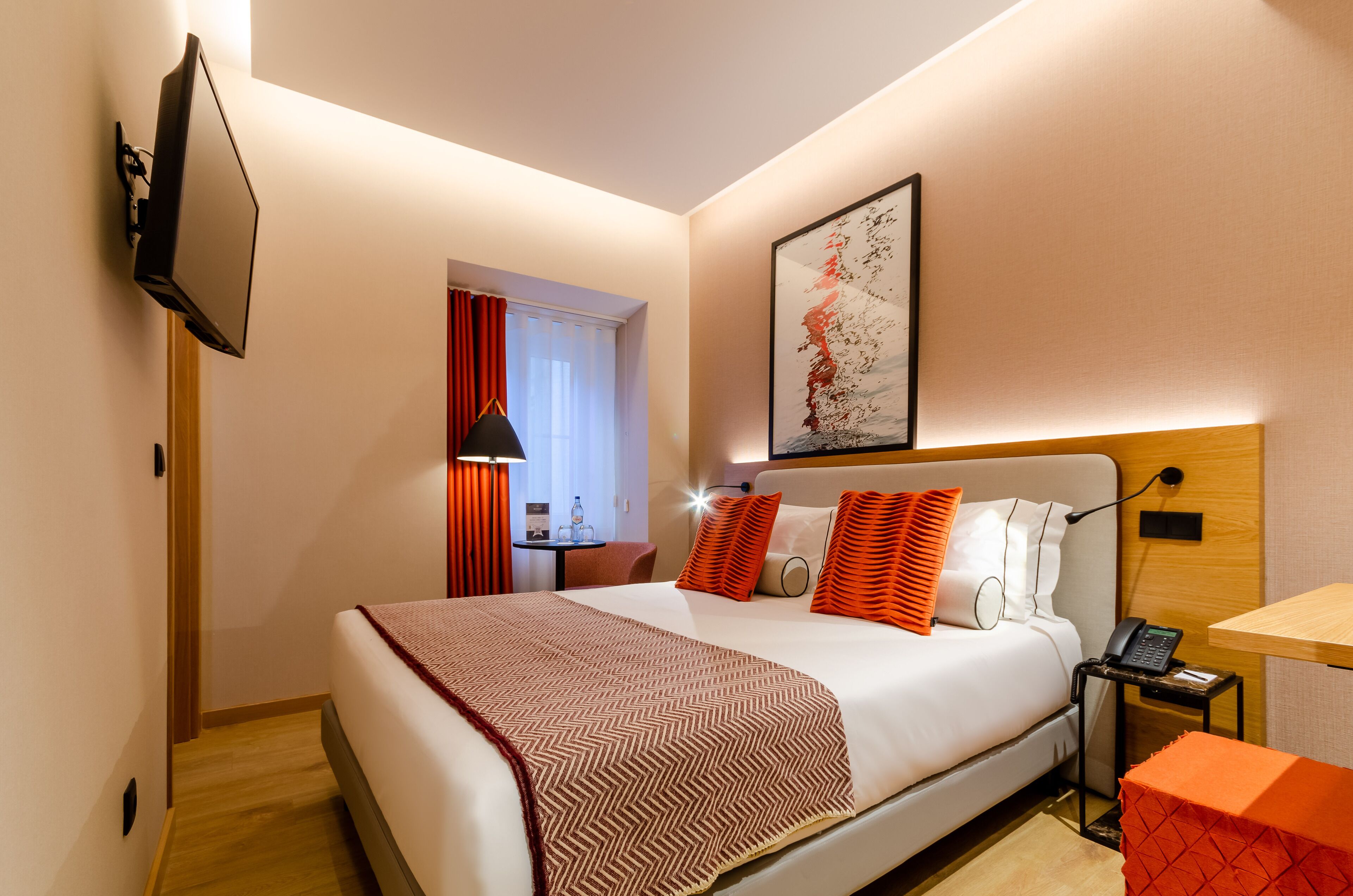 Una habitación de hotel con una cama, una mesita de noche con un teléfono, un televisor montado en la pared y un cuadro.