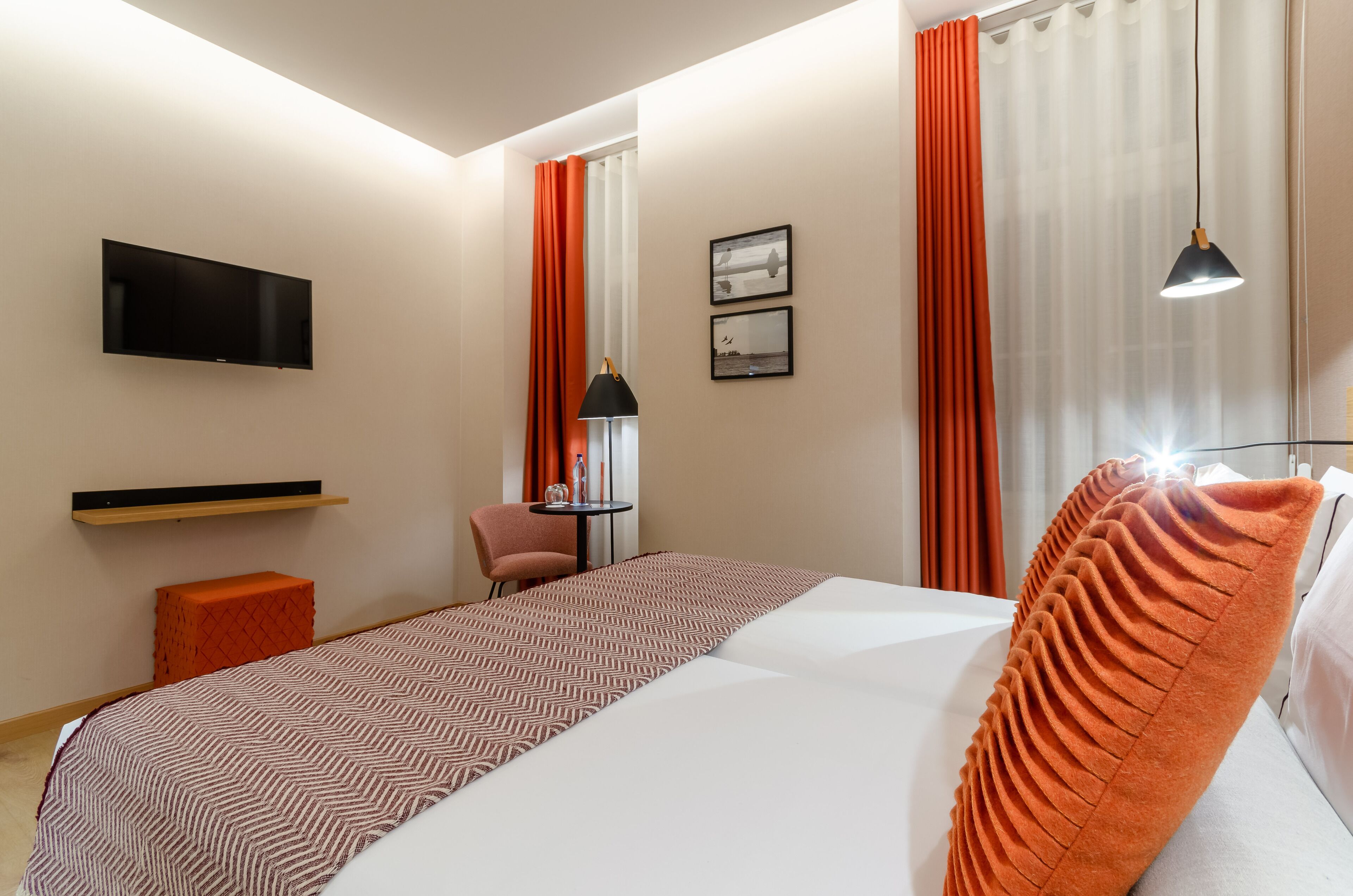 A hotel room with a bed, a TV mounted on the wall, a small table with a chair, and a window with curtains.