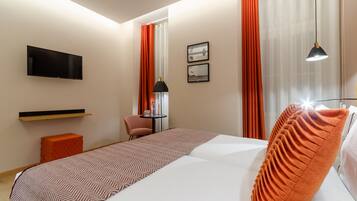 A hotel room with a bed, a TV mounted on the wall, a small table with a chair, and a window with curtains.