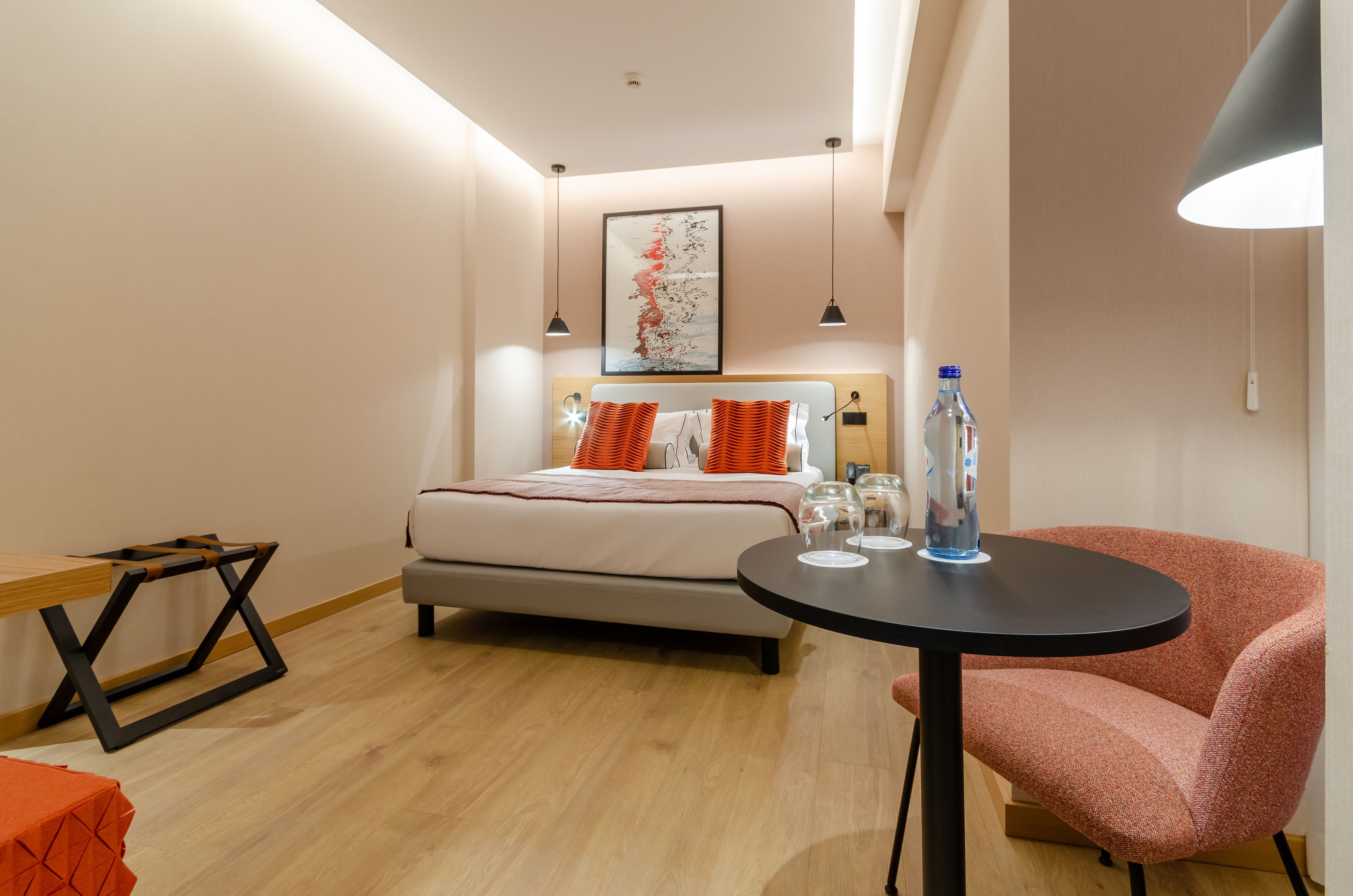 Une chambre d’hôtel moderne, équipée d’un lit, d’une table ronde, d’une chaise et d’une table de chevet sur laquelle se trouvent une bouteille et des verres.