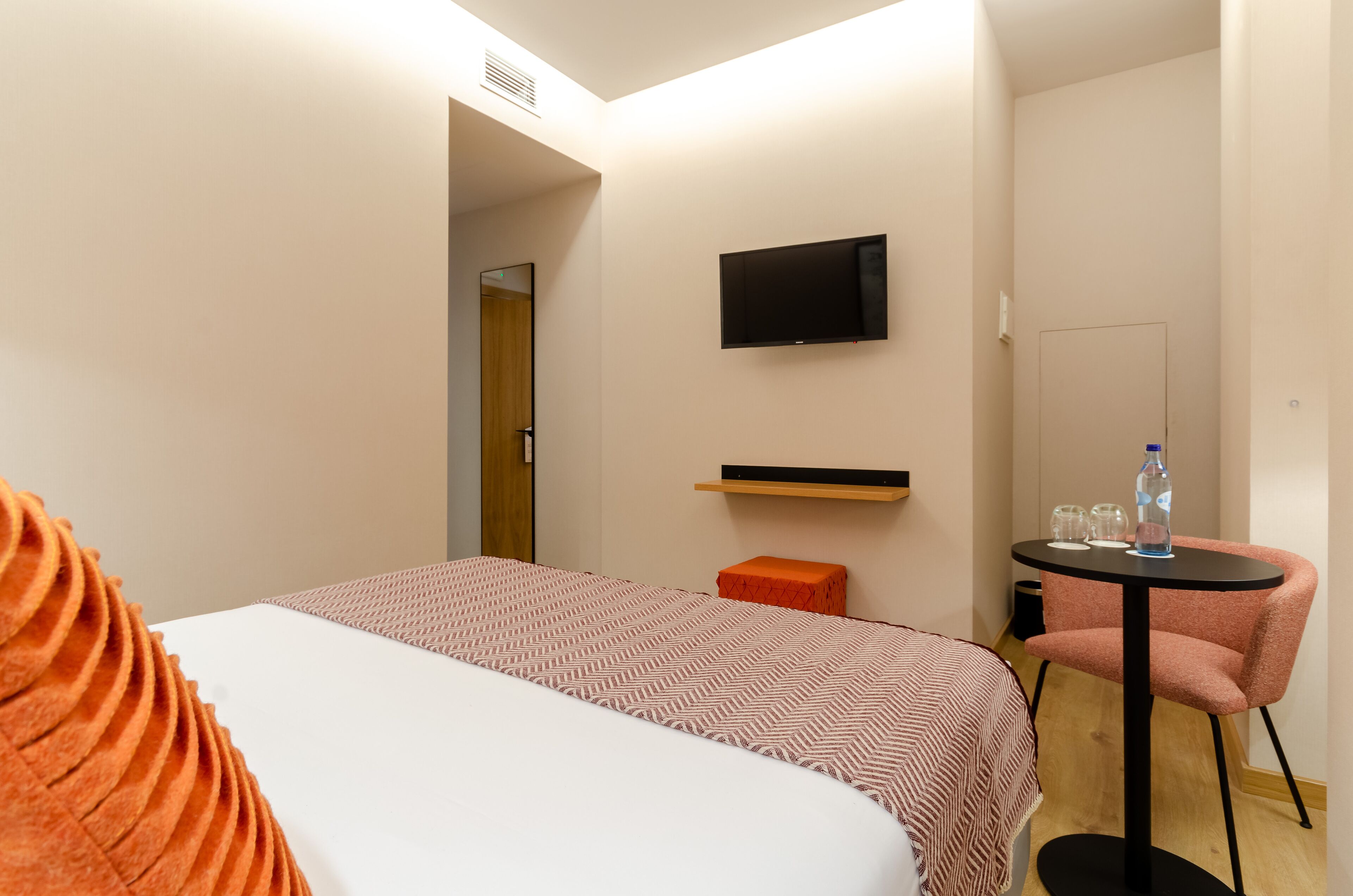 A hotel room with a bed, a television mounted on the wall, a small table with a chair, and a door leading to another room.