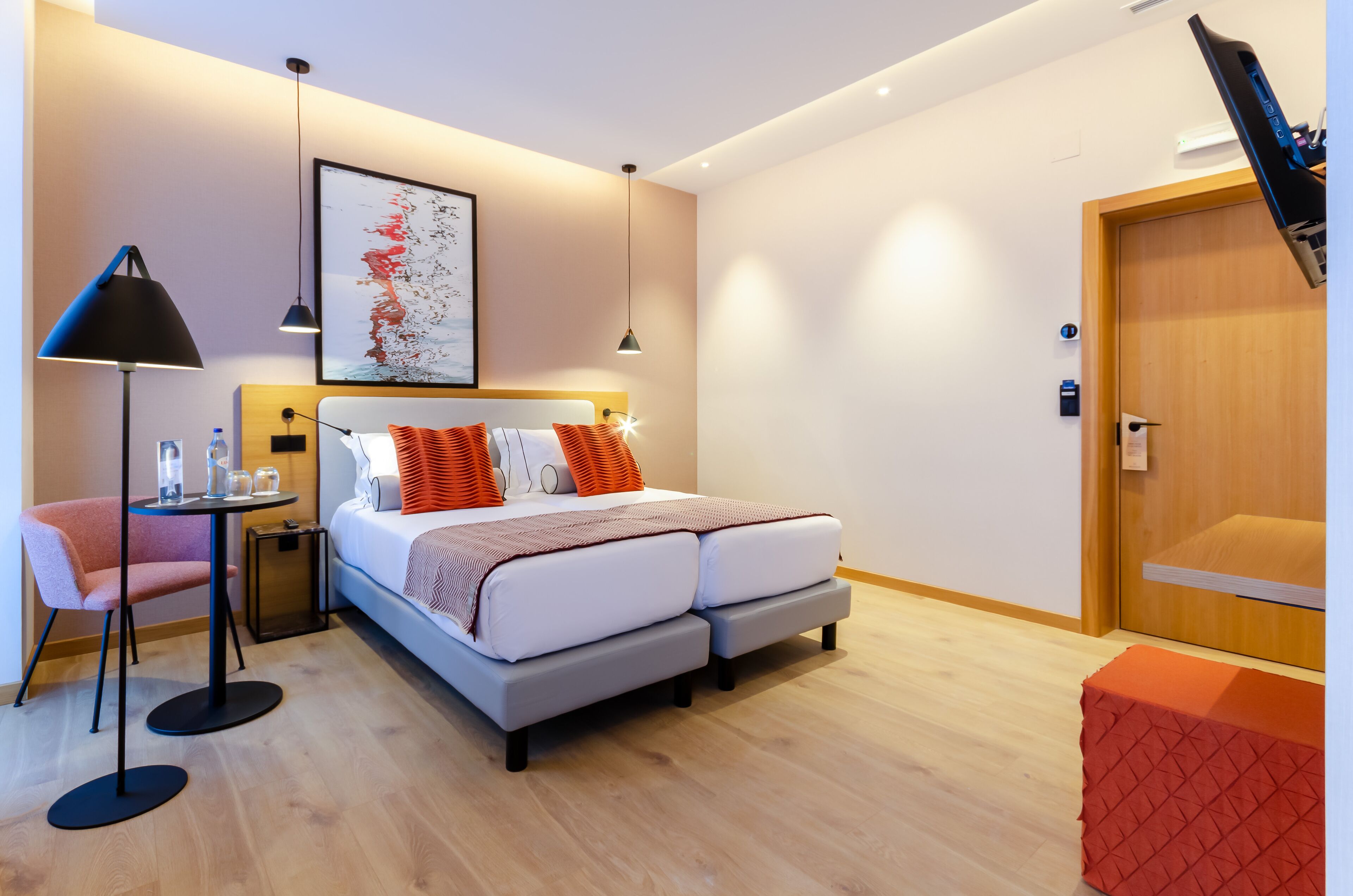 Quarto de hotel moderno com cama, escrivaninha, cadeira, um banquinho vermelho e porta de madeira.