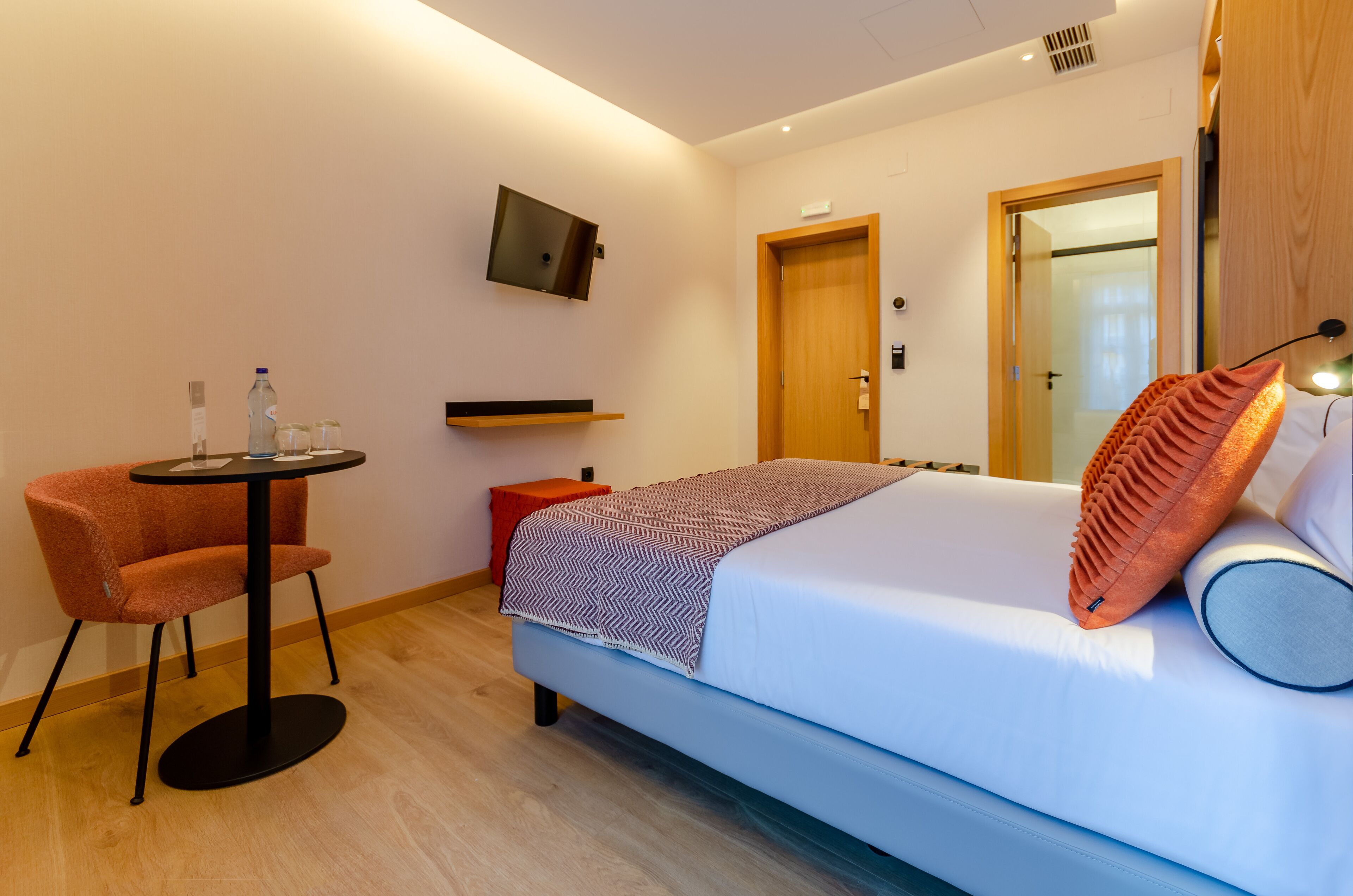 A hotel room with a bed, a small table, a chair, a TV mounted on the wall, and a wooden door.
