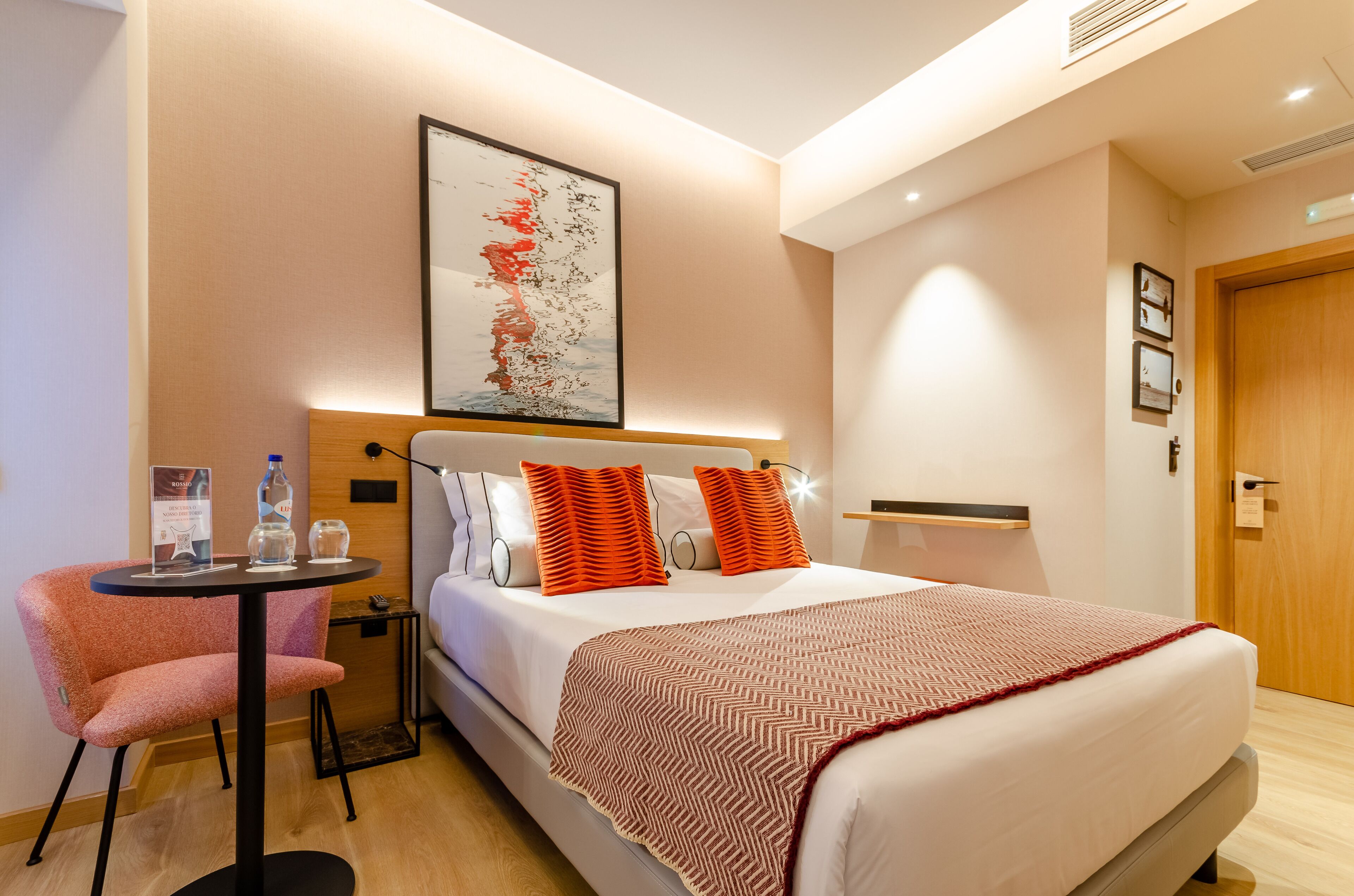 A hotel room with a bed, a chair, a table with water bottles, a framed picture on the wall, and a door.