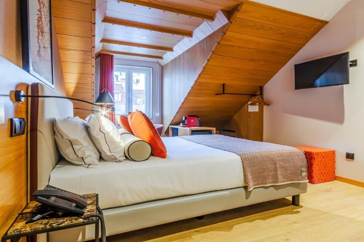 Un dormitorio con cama, mesita de noche con lámpara, televisor montado en la pared y ventana con cortinas.