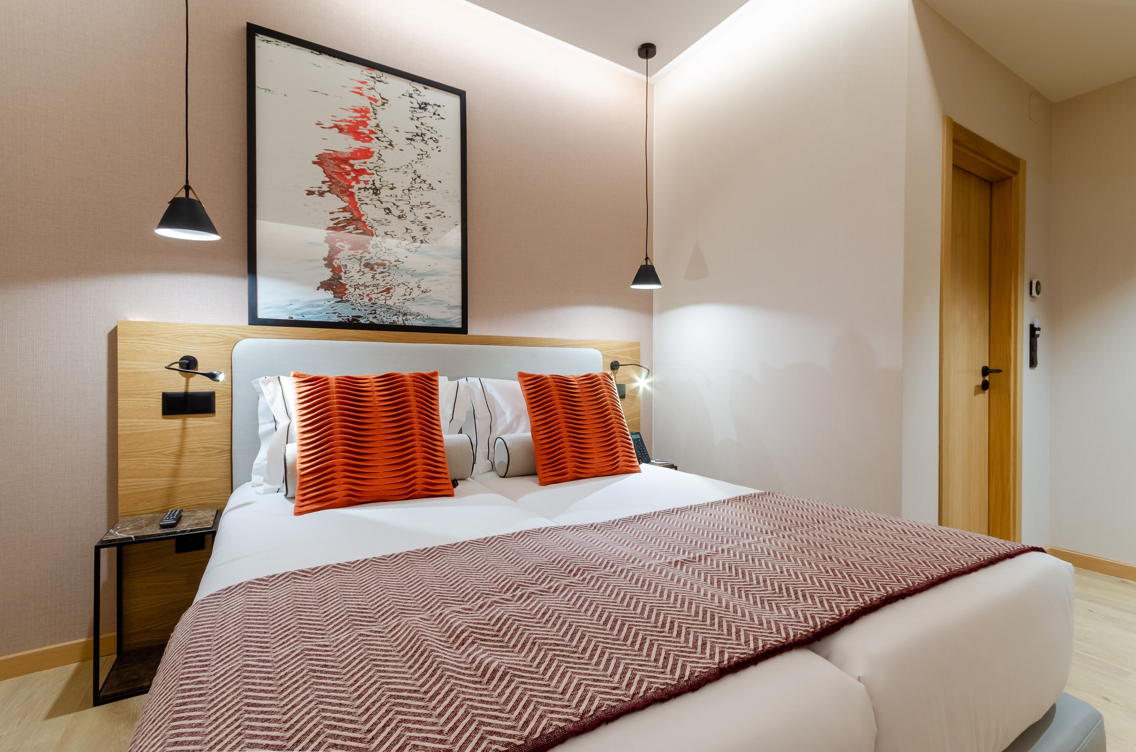Een moderne hotelkamer met een groot bed, twee nachtkastjes, een houten hoofdbord, een groot abstract schilderij en twee hanglampen.