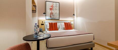 Una habitación de hotel moderna con una cama, una mesita redonda pequeña con una botella y vasos, dos sillas y un cuadro en la pared.