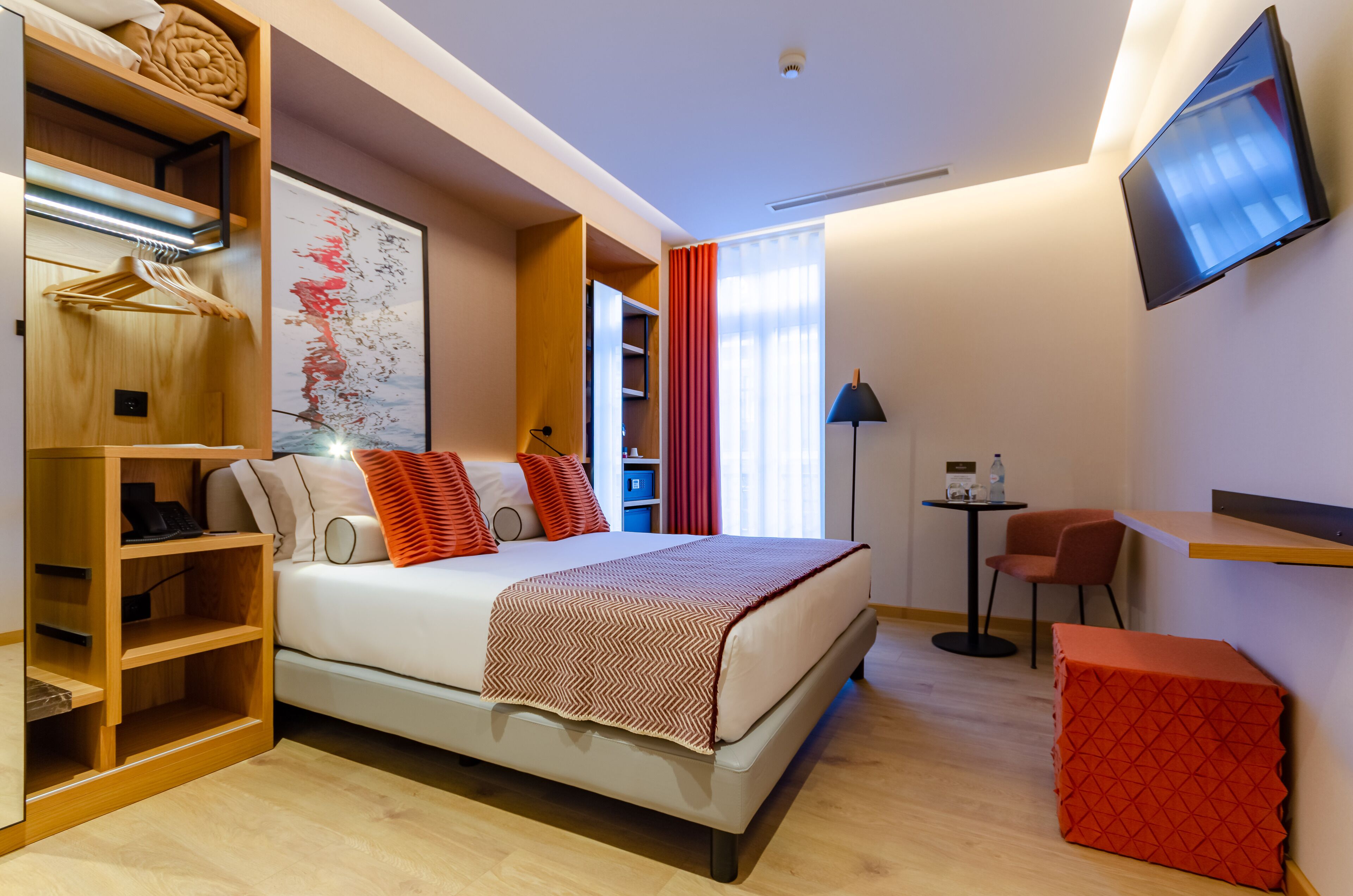 Une chambre d’hôtel moderne équipée d’un lit, d’une armoire, d’un bureau, d’une chaise et d’un téléviseur fixé au mur.