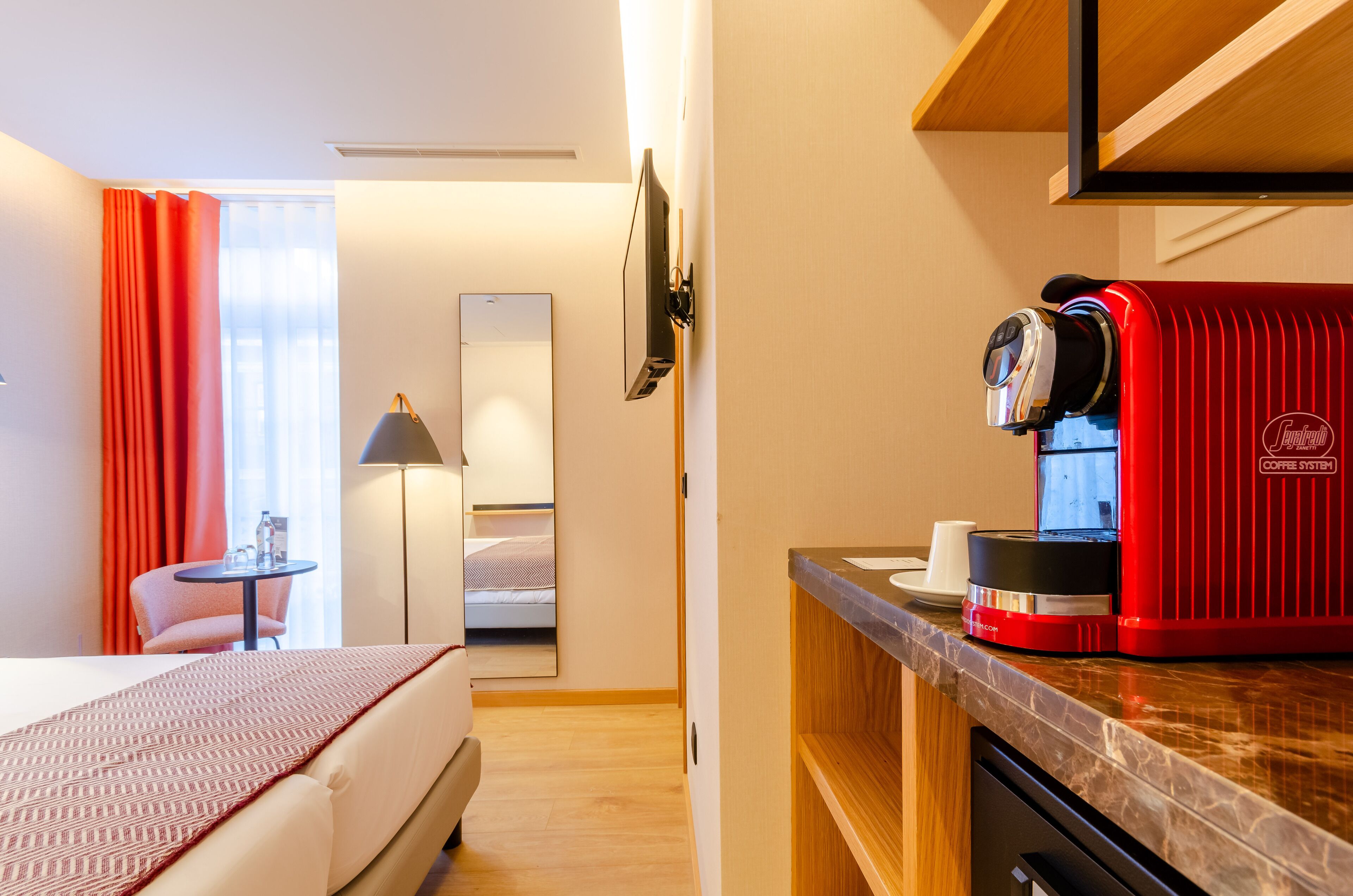 Ein Hotelzimmer mit einem Bett, einem kleinen Tisch mit Lampe, einer roten Kaffeemaschine und einem Spiegel an der Wand.