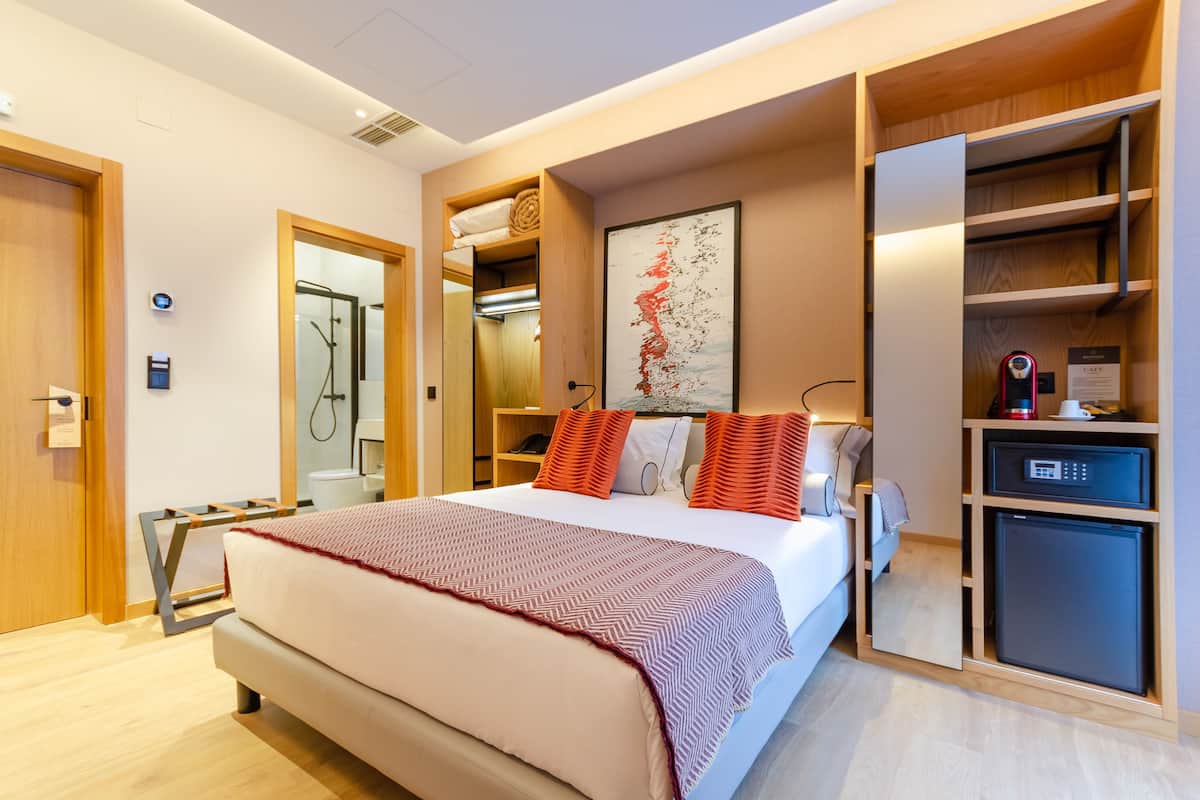 A modern hotel room with a large bed, a built-in wardrobe, a small refrigerator, and a shower area.