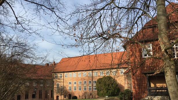 A large brick building with multiple windows and a red roof, surrounded by trees and a pathway.