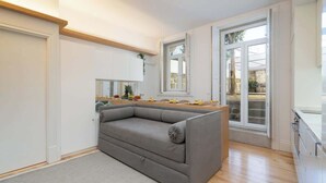 A modern living room with a grey sofa, wooden flooring, and large windows offering a view of the outdoors.