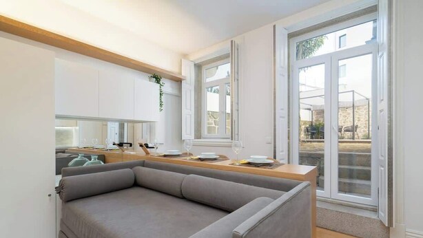 A modern living room with a grey sofa, a wooden dining table set for two, and a kitchen area with white cabinets and a window.