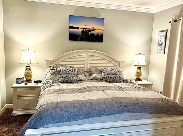 A bedroom with a bed, two bedside tables with lamps, a nightstand with a clock, and a framed picture on the wall. - XPLORIE ACTIVITIES & LINENS INCLUDED*!  Close to shopping, restaurants, community center, park & more! (Ocean City)