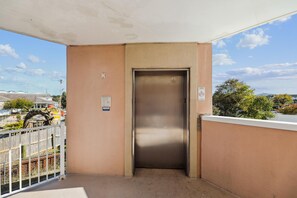Une porte de monte-charge fermée sur un balcon doté d’un sol en béton, d’une rambarde métallique et, en arrière-plan, d’un ciel dégagé.