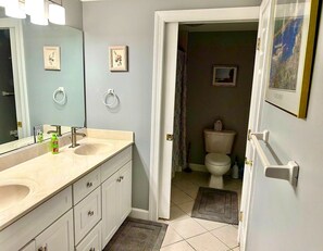 A bathroom with a double sink, a toilet, and a shower area. There are towels hanging on the wall and a rug on the floor. - XPLORIE ACTIVITIES & LINENS INCLUDED*!  Close to shopping, restaurants, community center, park & more! (Ocean City)