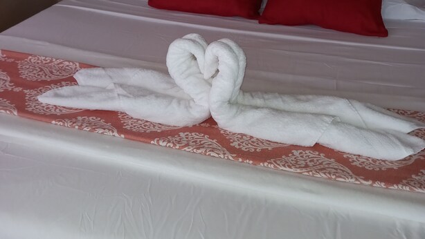 Un lit bien fait, recouvert d’une courtepointe blanche, avec deux oreillers rouges et une serviette disposée en forme de cœur sur le lit.