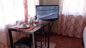 Un coin repas avec une table dressée pour deux personnes, un téléviseur posé sur un meuble et des rideaux à motifs.