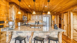 A wooden cabin interior with a kitchen featuring granite countertops, a central island with stools, and a stone accent wall. - Endless Views, Secluded Cabin Hot Tub & Fireplaces (Blue Ridge)