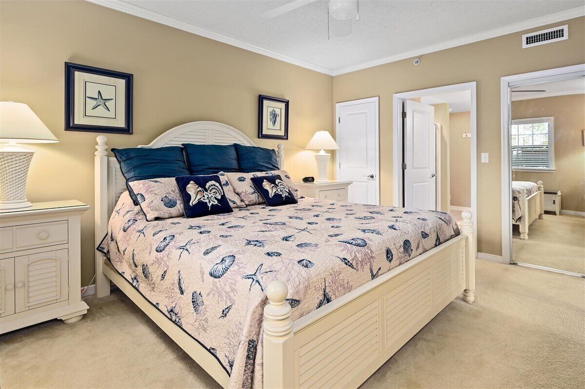 A bedroom with a large bed, two bedside tables with lamps, a dresser, and a door leading to another room.