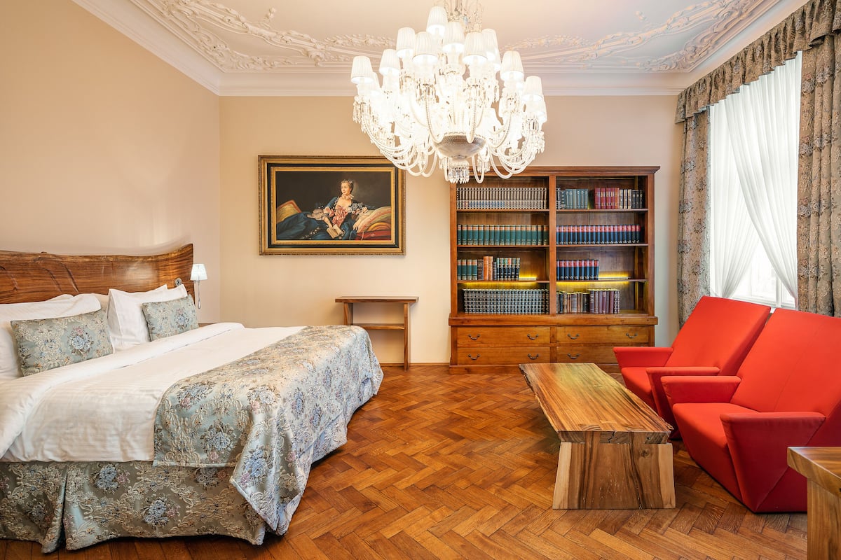 Habitación de hotel con una cama grande, una mesa de centro de madera, un sillón rojo, una estantería y una araña.