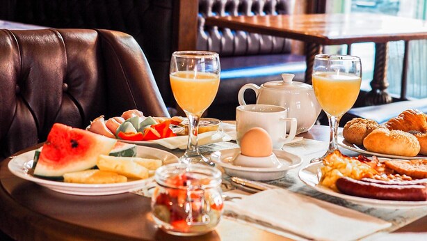 Una mesa de comedor con alimentos para el desayuno, como tostadas, huevos y pasteles, en una habitación con muebles de madera y vistas al exterior.