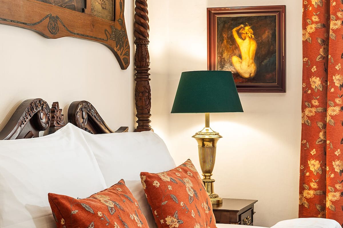 Una cama bien hecha con sábanas blancas y dos almohadas rojas con estampado floral, un cabecero de madera con detalles tallados, una lámpara de mesa verde y un cuadro enmarcado sobre la cama.