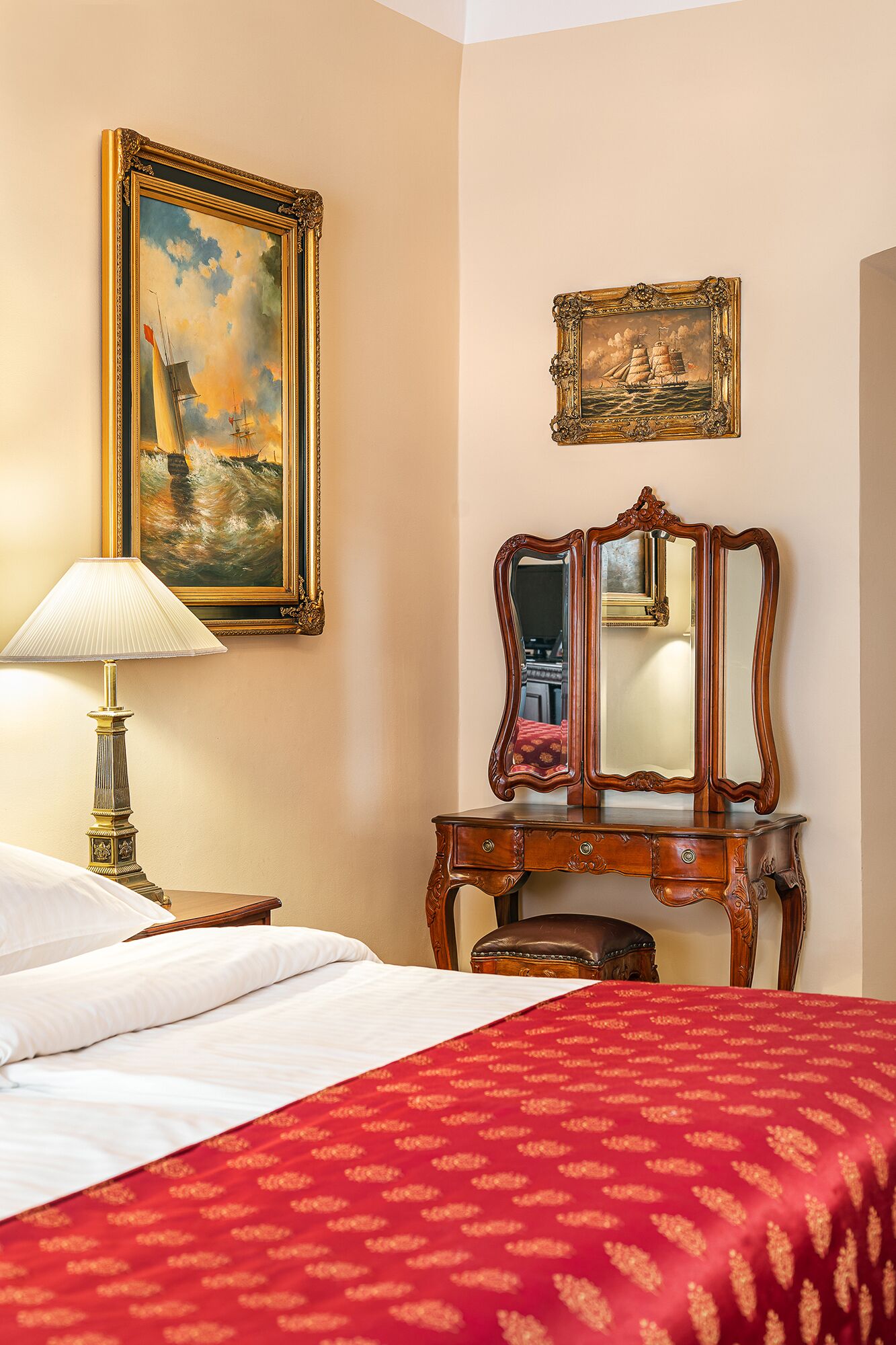 A hotel room with a bed, a nightstand, a lamp, a dresser with a mirror, and two paintings on the wall.