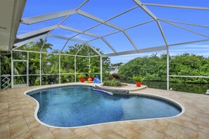 Una piscina interior de borde curvo, rodeada por una zona cubierta con estructura metálica y paredes de vidrio, que ofrece vistas a la vegetación y un cuerpo de agua al exterior.