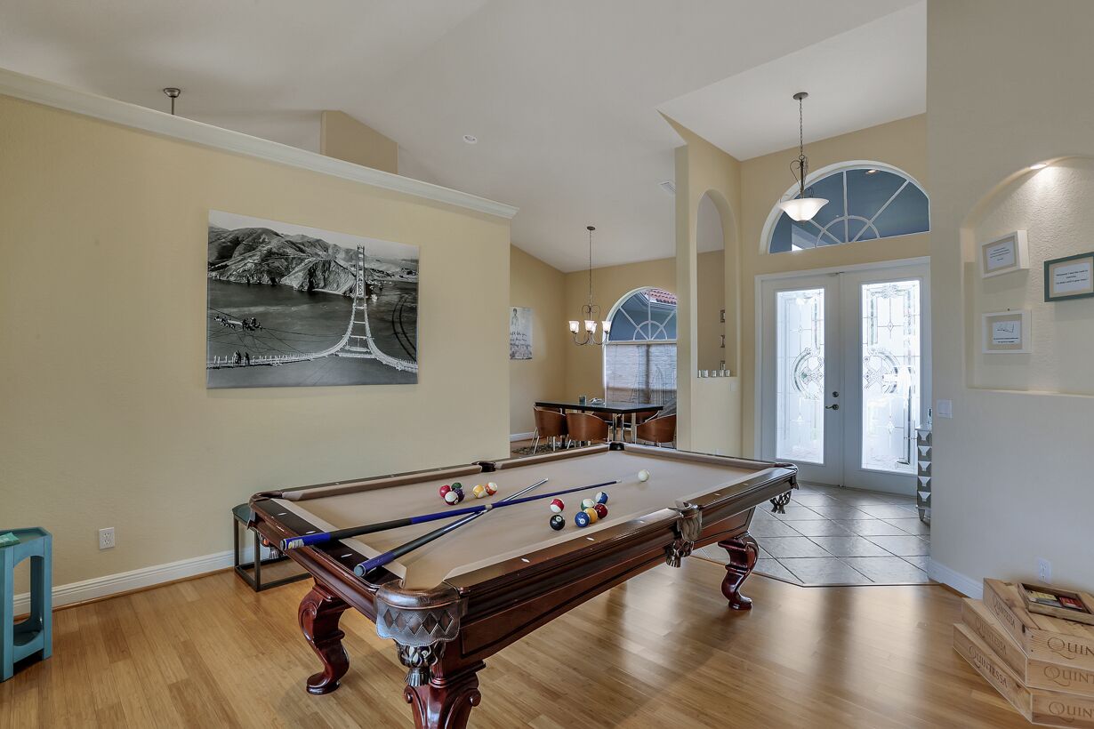 A pool table in a room with a large painting of a bridge, a dining area, and a staircase leading to a second floor.