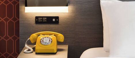 Une chambre d’hôtel avec une tête de lit en bois, une table de chevet sur laquelle se trouve un téléphone à cadran jaune et une horloge numérique.