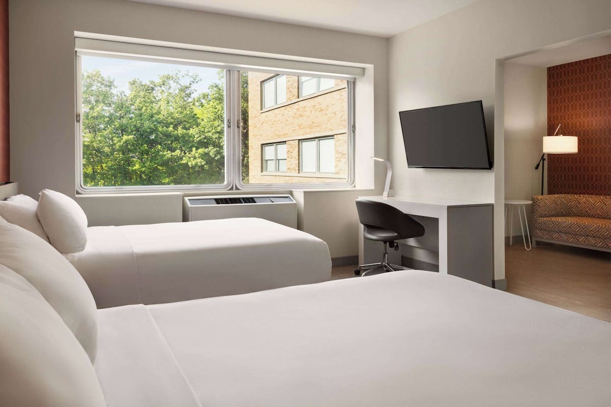 Une chambre d’hôtel moderne équipée de deux lits, d’un bureau, d’une chaise, d’une télévision et d’une fenêtre donnant sur des arbres et des immeubles.