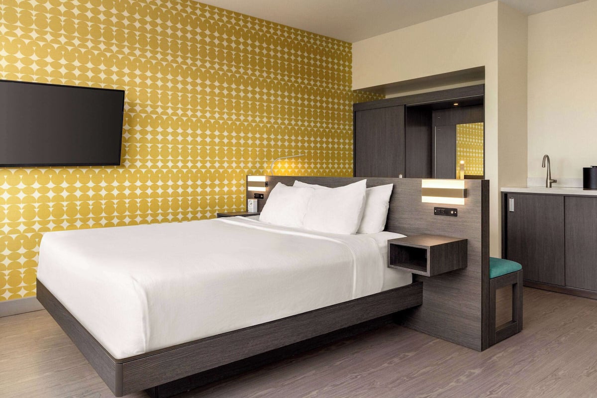 Une chambre d’hôtel moderne dotée d’un grand lit, d’une table de chevet avec une lampe, d’une armoire intégrée et d’un petit coin salon.