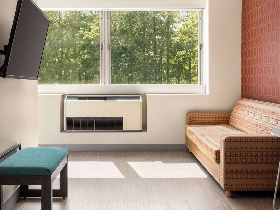 Una habitación con ventana, televisor de pared, sofá, mesa pequeña y un banco.