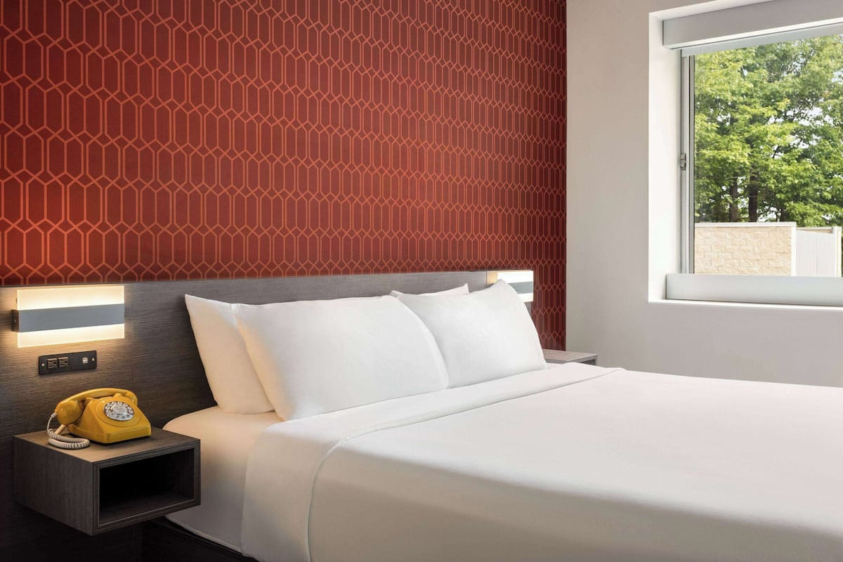 Une chambre d’hôtel avec un lit, une table de chevet sur laquelle se trouve un téléphone à cadran jaune, une lampe fixée au mur et une fenêtre donnant sur des arbres.