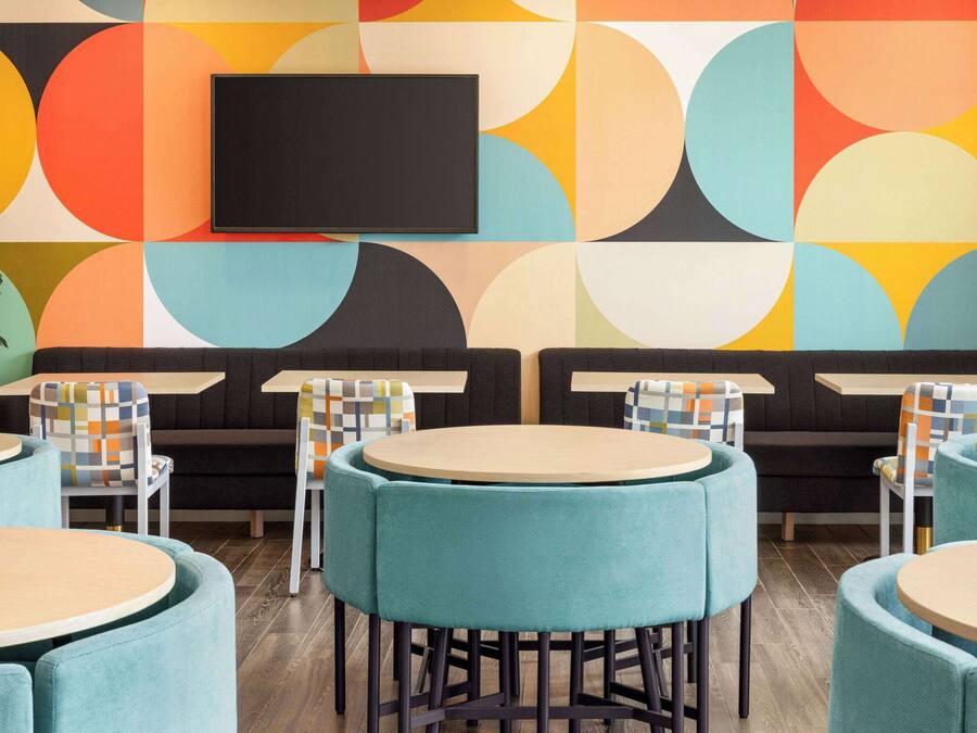 Un interior moderno con mesas redondas y sillas turquesa, un televisor de pantalla plana montado en la pared y un patrón geométrico colorido en la pared.