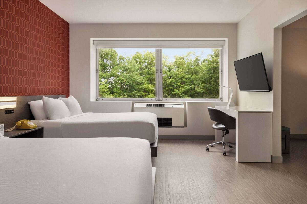 Une chambre d’hôtel avec deux lits, un bureau, une chaise, une télévision et une fenêtre donnant sur des arbres.