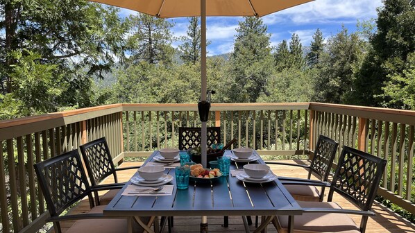 Una terraza de madera con una mesa lista para una comida, rodeada de sillas y con vistas a un área boscosa.
