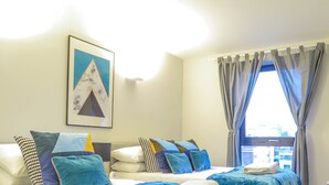 Una habitación de hotel con cama, mesita de noche, ventana con cortinas e imagen en un marco en la pared.