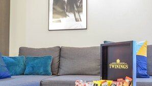 Una sala de estar con un sofá gris, cojines azules, un cuadro enmarcado en la pared y una caja de té Twinings sobre una mesa.