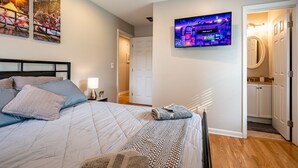 A bedroom with a bed, pillows, a bedside table with a lamp, a TV on the wall, and a door leading to a bathroom. - Discover Your Beautiful Dream Vacation Rental at The Battery - Truist Park!  (Smyrna)
