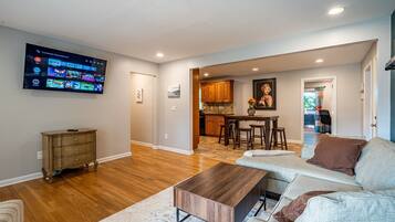A modern living room with a flat-screen TV, wooden flooring, a sofa, a coffee table, and a kitchen area with wooden cabinets and a dining table.